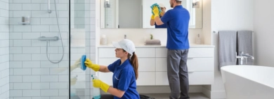 A two-person cleaning team performing a detailed Bathroom service in a modern, pristine bathroom, with one person cleaning a glass shower door and the other polishing a large mirror above a double vanity.
