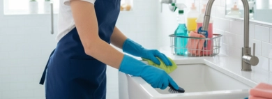 A smiling cleaner, wearing a blue apron and gloves, meticulously scrubs a white bathroom sink, representing professional bathrooms cleaning (residential) services.