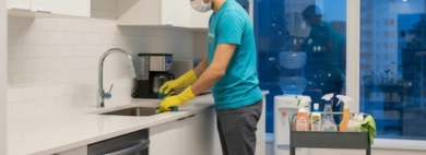 A uniformed cleaner in a mask scrubs the sink as part of a professional service to clean kitchen areas in a modern office breakroom at night.