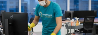 A professional cleaner wearing a mask provides a thorough service for cleaning desktops, carefully disinfecting an office workstation with spray and a cloth.