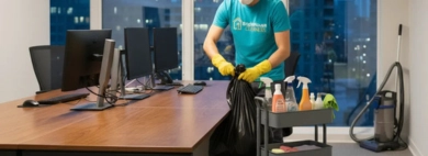 A professional cleaner in a mask and gloves handles the disposal of all trash by emptying office wastebaskets into a large bag after hours.