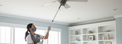 A professional cleaner on a ladder, diligently cleaning a ceiling fan with an extendable duster in a spacious, tidy living room, demonstrating thorough Dust Ceiling Fans Cleaning services.