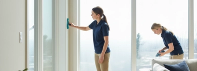 Two professional technicians in uniform perform interior cleaning inside a bright, modern living room, meticulously cleaning large windows with squeegees.