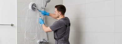 A cleaner in blue gloves and grey overalls thoroughly scrubs a shower wall with a brush, with a freestanding tub nearby, depicting professional Shower and Tub Scrubbing.