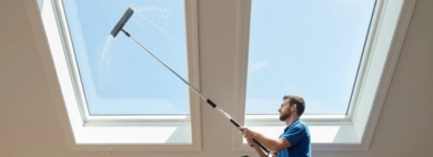 Two professional technicians are performing skylight cleaning inside a home. One stands on a tall ladder using a long-handled squeegee on an upper skylight, while the other cleans a lower skylight with a cloth, ensuring bright, clear views.