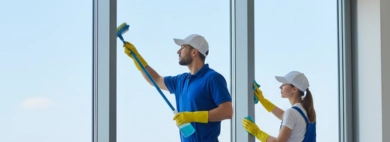 A professional two-person cleaning team, dressed in blue uniforms and yellow gloves, expertly performing a Window service on large, expansive windows overlooking a city skyline, using squeegees and cleaning solutions.