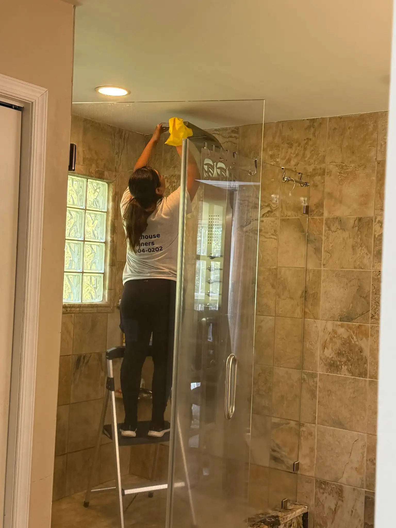 A Brighthouse Cleaners on a stepladder provides a deep cleaning service, meticulously cleaning a glass shower door.