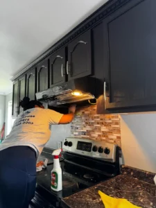 A Brighthouse Cleaners professional delivering a detailed kitchen cleaning service, focused on cleaning a range hood over a stove with black cabinets.