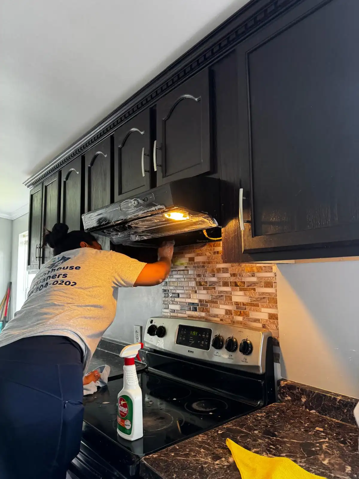 A Brighthouse Cleaners professional delivering a detailed kitchen cleaning service, focused on cleaning a range hood over a stove with black cabinets.